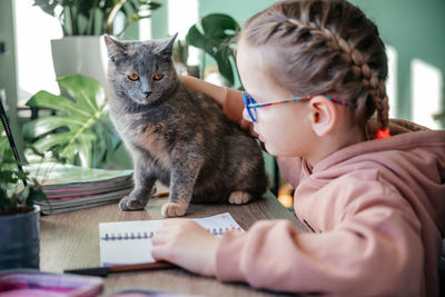 Little girl in eyeglasses with her pet cat having online lesson during covid lockdown, distance