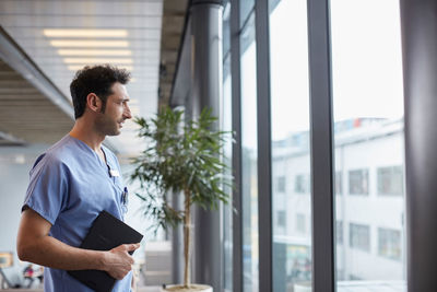 Thoughtful young male nurse while looking through window while standing at corridor in hospital