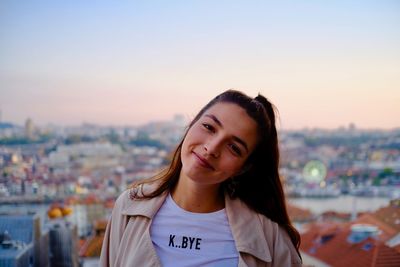 Portrait of smiling young woman standing in city against sky during sunset