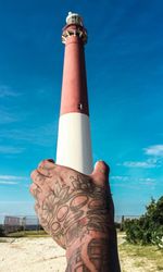 Optical illusion of cropped hand holding lighthouse against blue sky at beach 