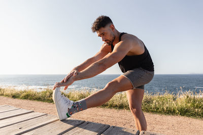 Side view of muscular male doing forward bends and warming up before workout while standing near promenade on seashore