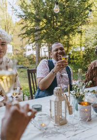 Happy gay man holding wineglass while sitting with friends during dinner party in back yard