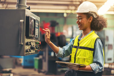 Standing in front of a control panel, a female industrial electrical engineer with a safety hardhat.