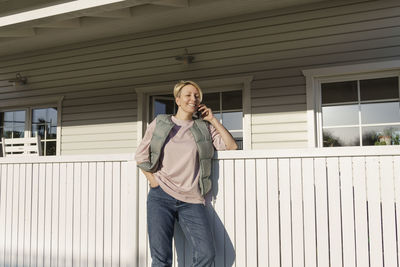 Woman talking on smart phone while leaning on railing outside house