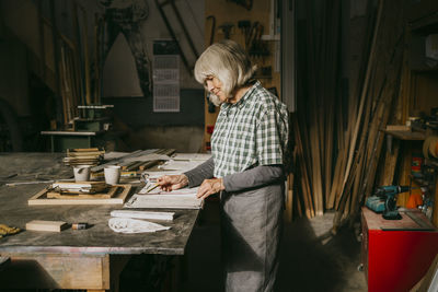 Side view of senior female carpenter reading document while standing at workshop