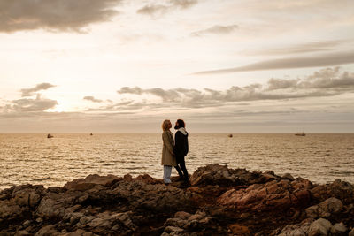 Boyfriend and girlfriend in outerwear looking at each other while standing on stones against rippling sea and cloudy sky in evening in aviles, spain