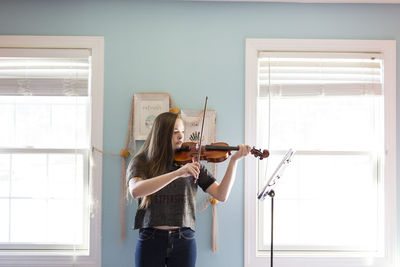 Woman playing violin while standing by window at home