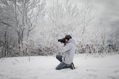 Side view of man photographing while kneeling on snow covered field