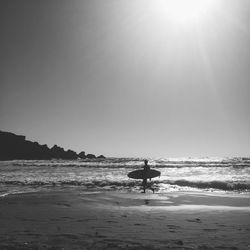 Surfer carrying surfboard on shore against clear sky