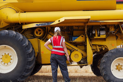 Back view of anonymous female worker in uniform examining combine harvester while working in agricultural field in countryside on summer day