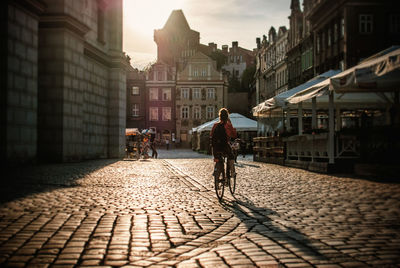 Rear view of woman cycling on cobbled street in city during sunset