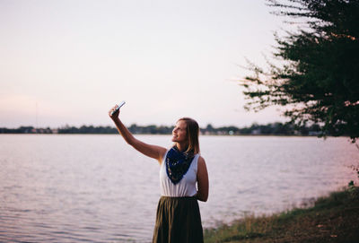 Woman taking selfie at lakeshore against sky during sunset