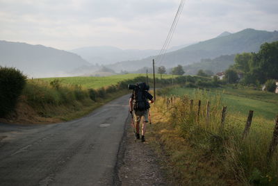 Rear view of hiker walking on roadside against sky