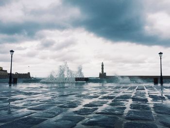 Waves splashing on promenade against cloudy sky