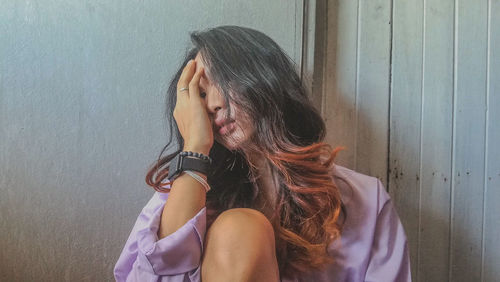 Woman with hand covering face sitting against wall at home