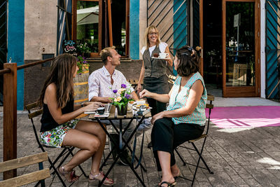 Side view of young women and man drinking coffee at table and communicating with waitress in outside cafe