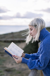 Side view of young female in blue sweater leaning on wooden fence and enjoying favorite poetry while spending time at seaside