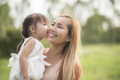 Cute daughter kissing on mother cheek against trees