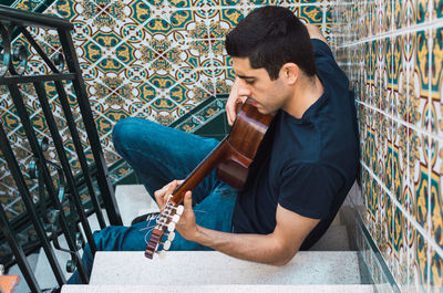 Young man playing guitar sitting on a staircase with beautiful tiles.