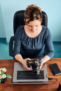 High angle view of woman holding camera sitting by laptop at home