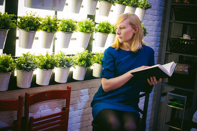 Young woman holding book against potted plants at greenhouse