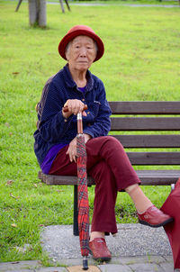 Portrait of senior woman with umbrella sitting on bench by grassy field at park