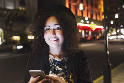 Portrait of smiling woman biting lip while holding smart phone in city at night