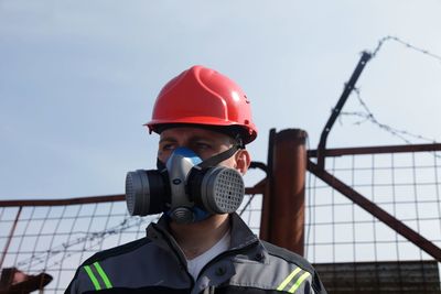 Close-up of worker wearing gas mask with fence in background against sky