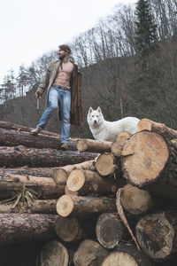 Low angle view of young man standing with dog on logs of wood in forest