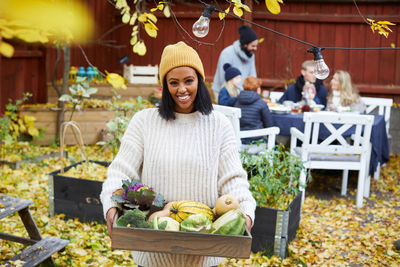 Portrait of happy woman standing with vegetable basket while family and friends sitting at table in yard