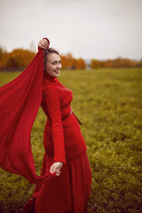 Woman in a red dress and sweater stands in a field with trees in autumn
