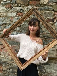 Portrait of young woman holding photo frame against wall