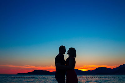 Couple standing on shore against sky during sunset