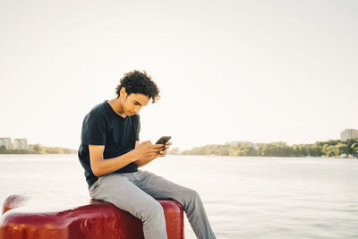 Teenage boy using mobile phone while sitting on bollard by river against clear sky during sunny day