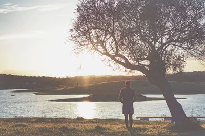 Rear view of man standing by lake against sky during sunset