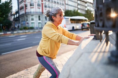 Elderly female athlete smiling and doing push ups near park fence during fitness workout in morning on street