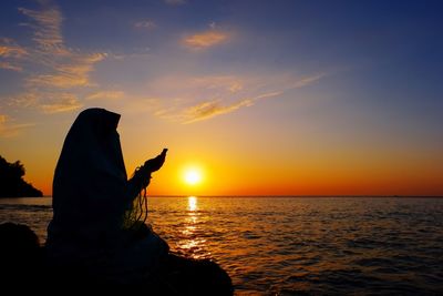 Silhouette woman praying by sea against sky during sunset