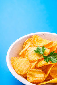 Fried corrugated golden potato chips with parsley leaf in wooden bowl on blue background