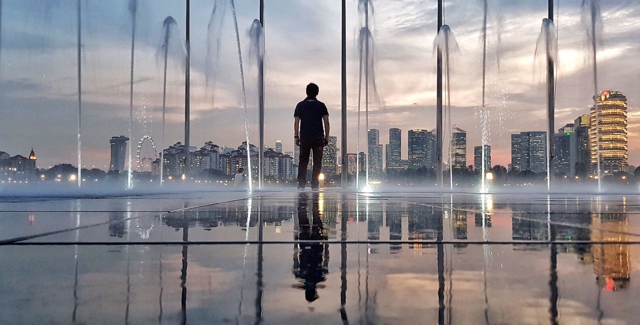 REFLECTION OF MAN STANDING ON WATER IN CITY