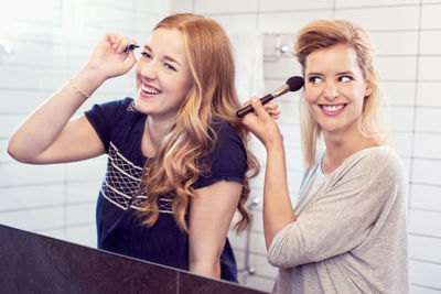Happy young sisters applying makeup in mirror