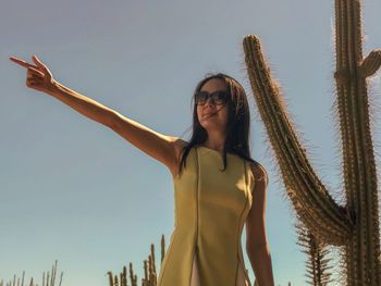 Low angle view of woman pointing against tall cactus and blue sky.