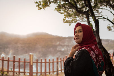 Portrait of young woman standing by tree against sky