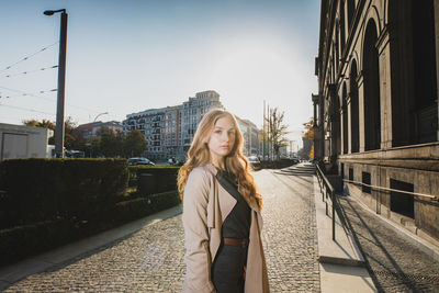 Portrait of young woman standing on footpath in city against sky
