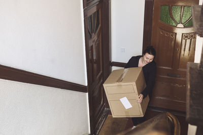 High angle view of smiling delivery woman with cardboard box standing against door