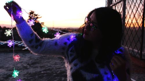 Young woman holding illuminated string lights during sunset