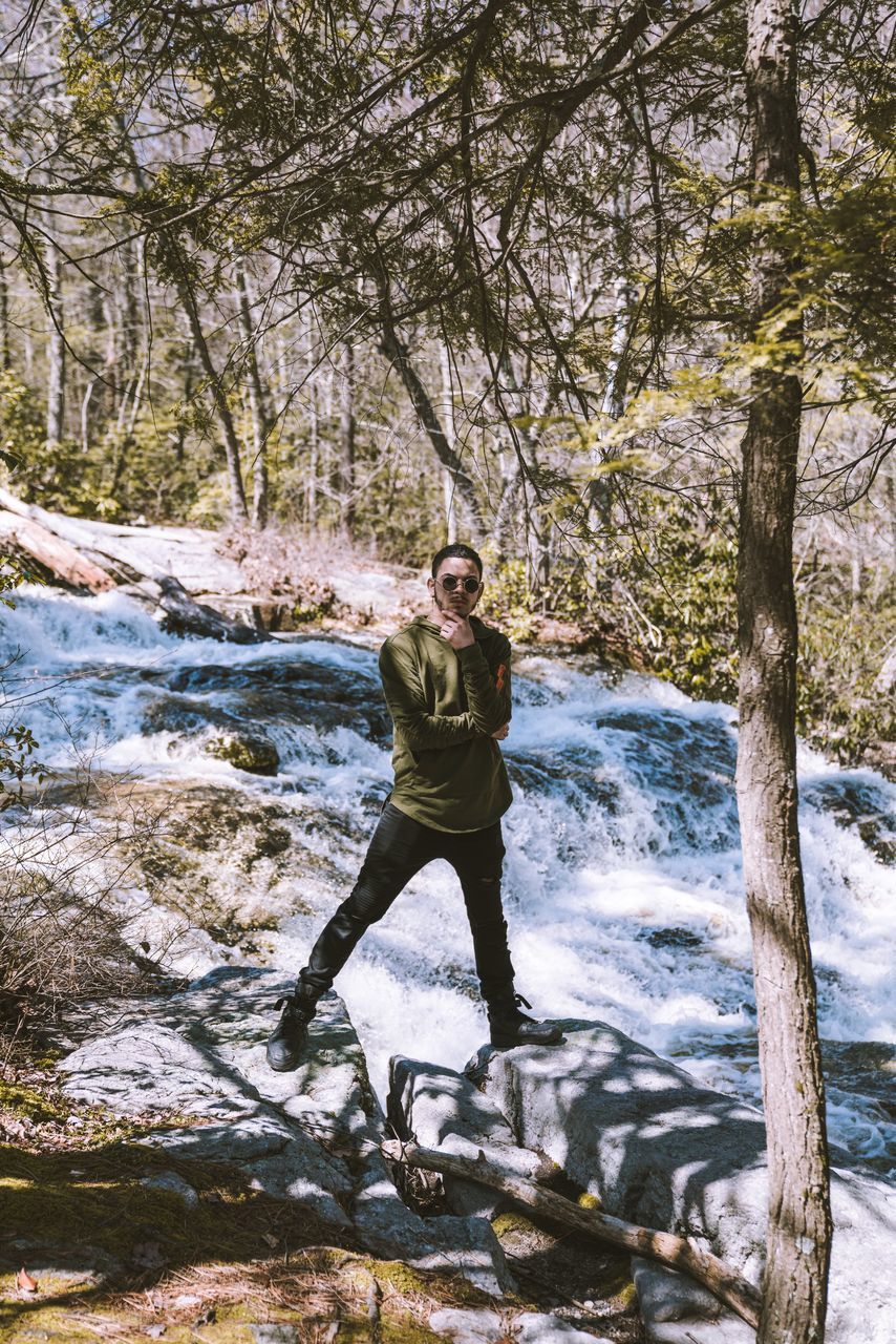 one person, tree, full length, water, plant, real people, young adult, lifestyles, casual clothing, front view, leisure activity, portrait, standing, nature, day, looking at camera, young men, forest, outdoors, flowing water, stream - flowing water