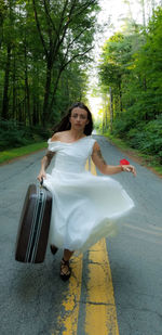 Woman holding suitcase while running on road amidst trees