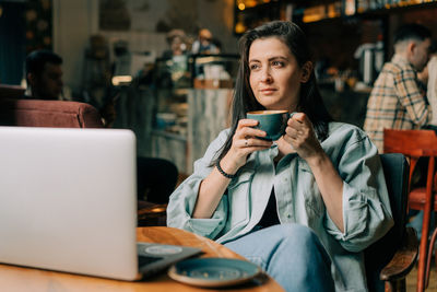 Young brunette woman sitting in a coffee shop drinking coffee and working on a laptop.