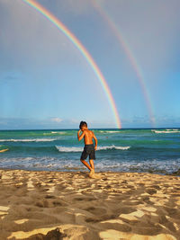 Shirtless boy walking against rainbow at beach