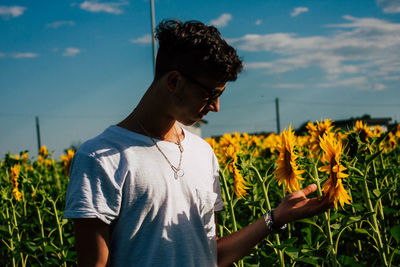 Young man touching sunflower on field against sky during sunny day
